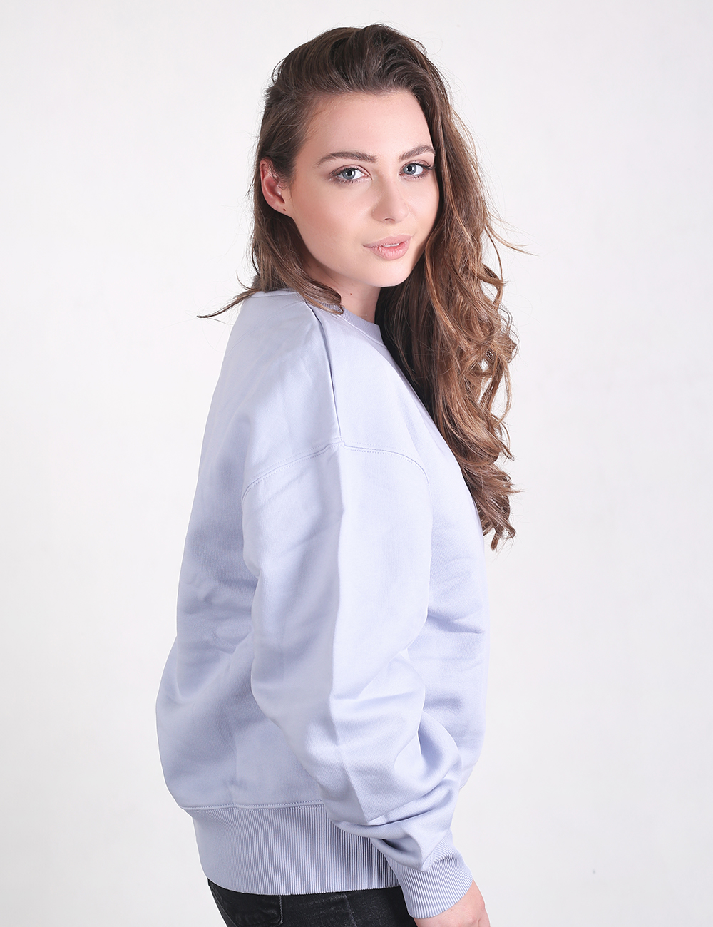 A woman with long, wavy brown hair looks over her shoulder while wearing an oversized, light pastel purple or lilac crewneck sweatshirt.