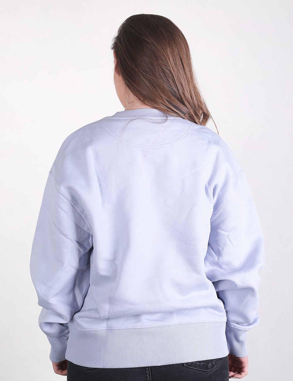 A person with long brown hair stands facing away from the camera, wearing an oversized, light purple crewneck sweatshirt and dark pants against a plain white background.