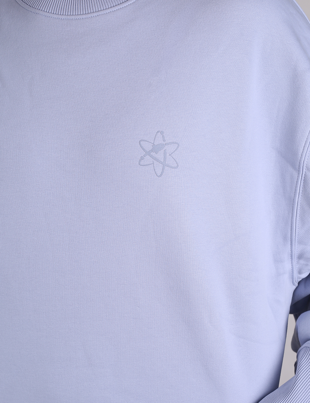 A close-up of a light purple or lavender ribbed knit sweatshirt or long-sleeve shirt.On the left chest area, there is a small, subtle embroidered or printed graphic depicting an atom symbol with a small, stylized shape integrated inside the center. The fabric shows a clear vertical ribbed texture throughout.