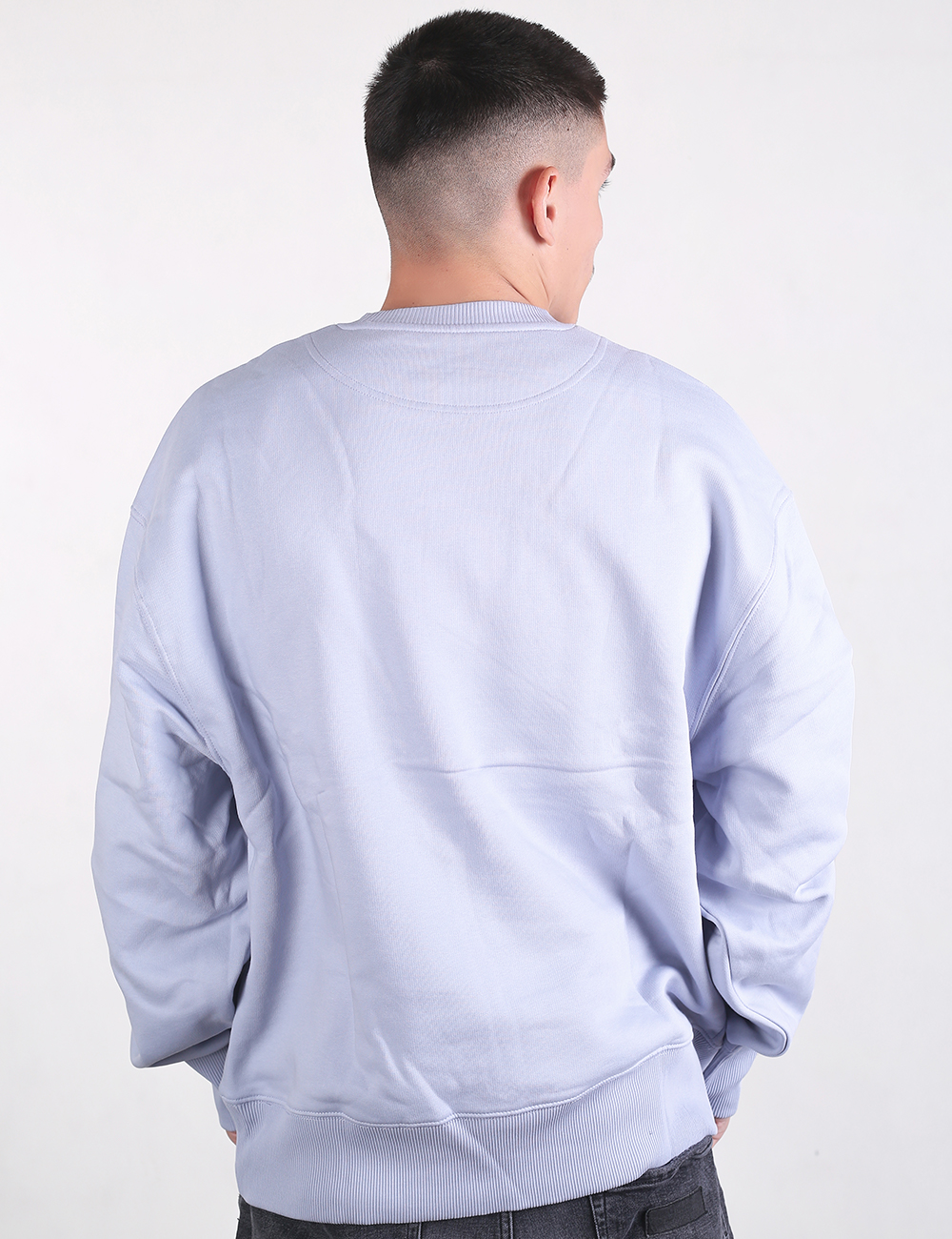 A man viewed from behind wearing a light lavender-blue, oversized crewneck sweatshirt. The sweatshirt features dropped shoulders and a thick ribbed waistband. He has short hair with a high fade haircut, and the waistband of dark grey jeans is visible at the bottom.