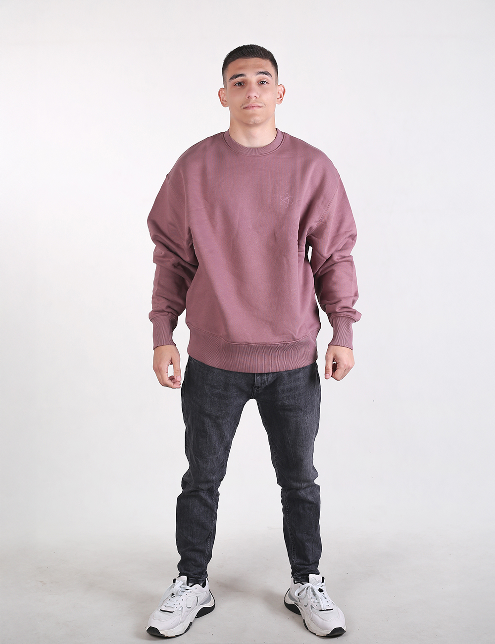 A full-length shot of a man standing against a plain white background, wearing a dusty rose or mauve oversized crewneck sweatshirt, dark grey slim-fit jeans, and white athletic sneakers.