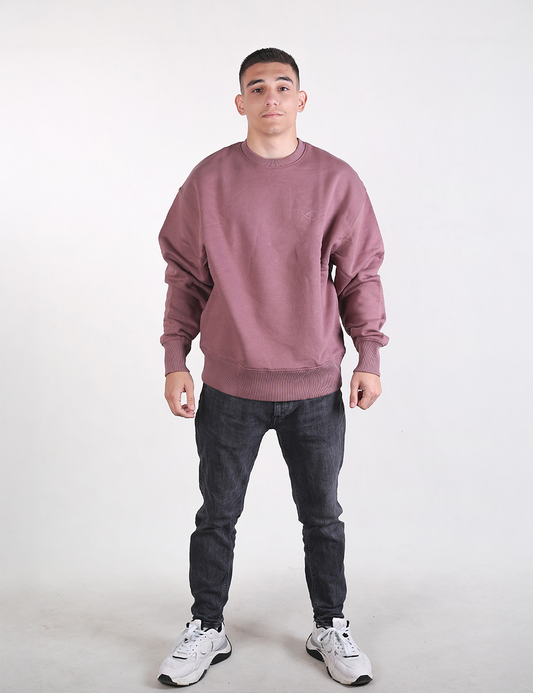 A full-length shot of a man standing against a plain white background, wearing a dusty rose or mauve oversized crewneck sweatshirt, dark grey slim-fit jeans, and white athletic sneakers.