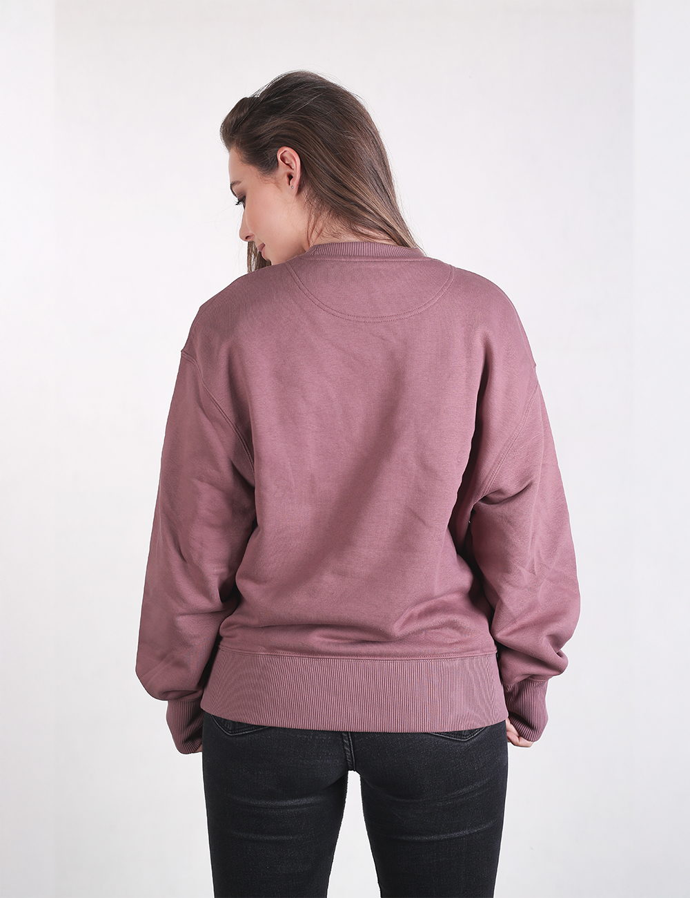 A woman with long brown hair stands facing away from the camera, wearing an oversized, mauve-colored crewneck sweatshirt and black pants.