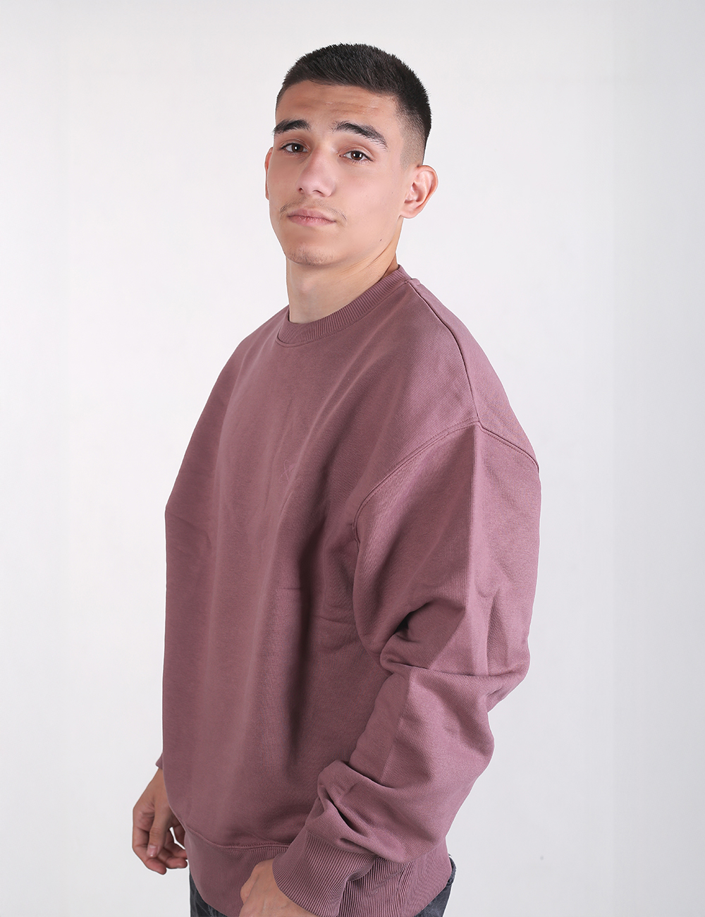 A three-quarter side profile shot of a young man with a buzz cut, modeling an oversized, solid mauve or dusty pink crewneck sweatshirt. The sweatshirt features dropped shoulders, long sleeves, and a classic ribbed neckline and hem. He is standing against a solid, light grey background with his hand partially in his pocket.
