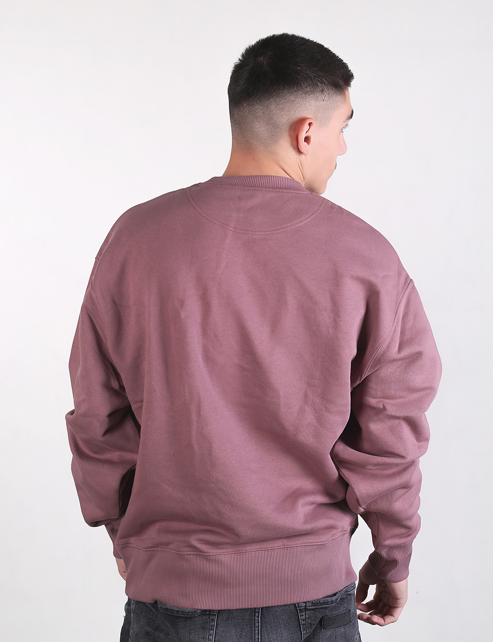 A back-facing view of a young man with a short fade haircut, wearing an oversized, dusty pink-purple crewneck sweatshirt and dark grey jeans against a plain white background.