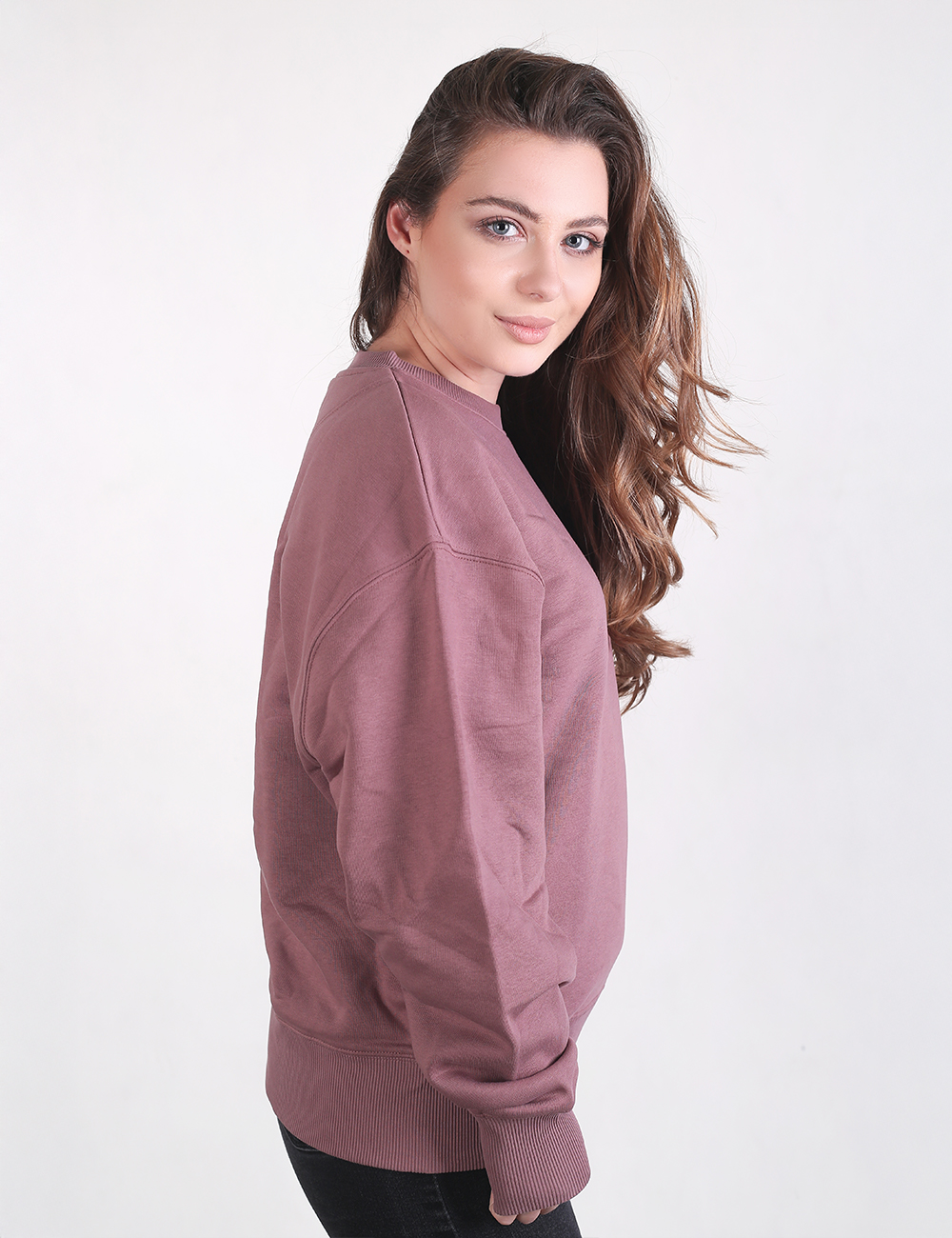 A young woman with long, wavy brown hair looks over her shoulder while wearing a mauve, oversized crewneck sweatshirt with ribbed cuffs.
