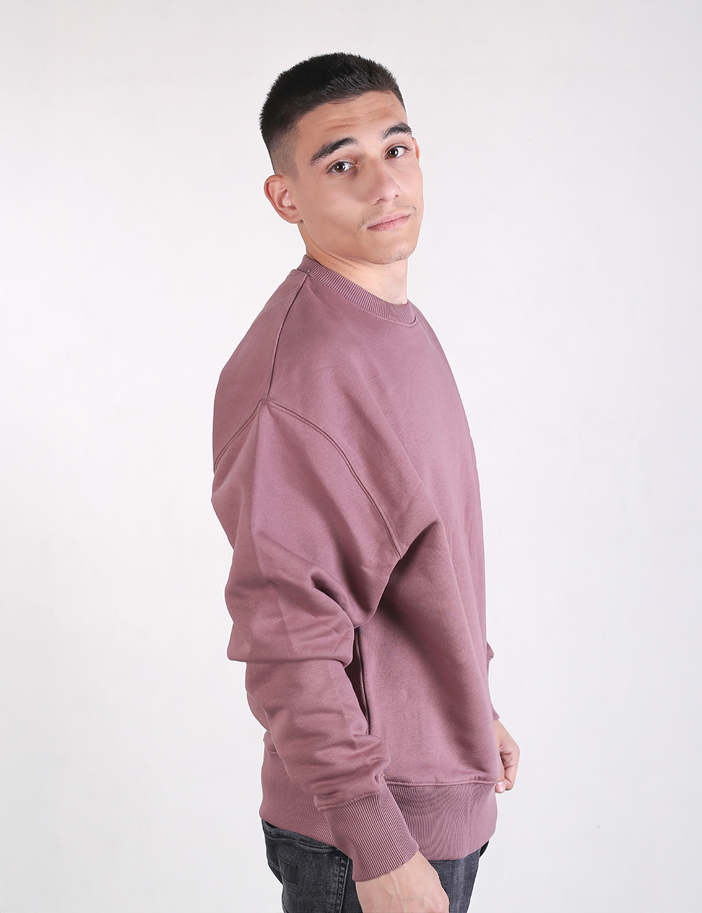 A man is pictured from the side against a plain white background, wearing an oversized, mauve-pink crewneck sweatshirt with dropped shoulders and ribbed detailing at the cuffs and hem, paired with dark jeans.