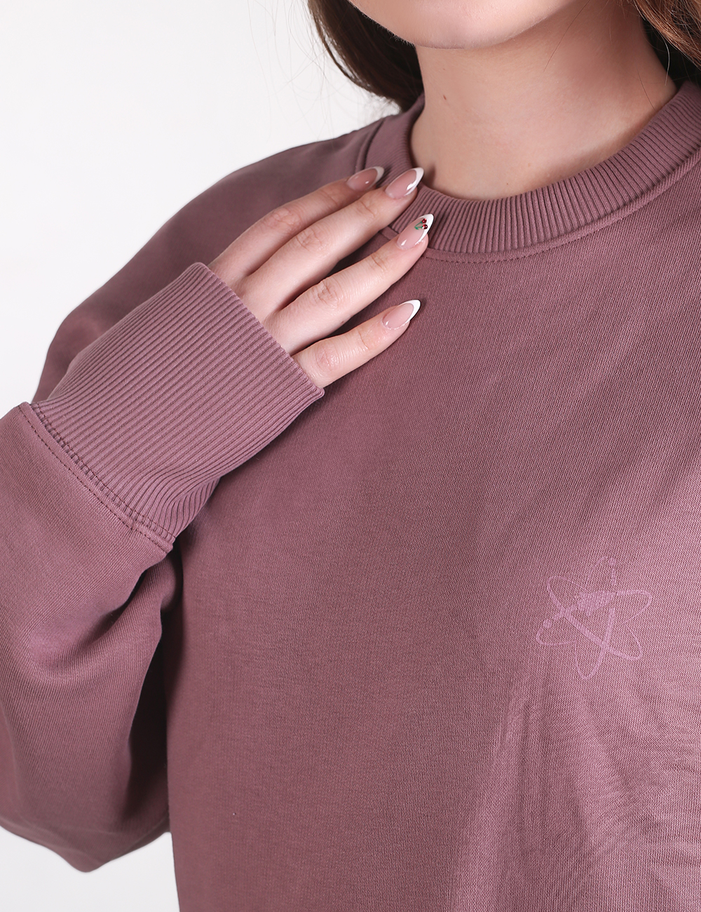 A close-up shot of a person wearing a mauve, long-sleeved crewneck sweatshirt with ribbed detailing on the cuffs and collar. Their right hand, featuring long manicured nails, rests gently near the neckline. A faint, tonal star logo is visible on the chest.