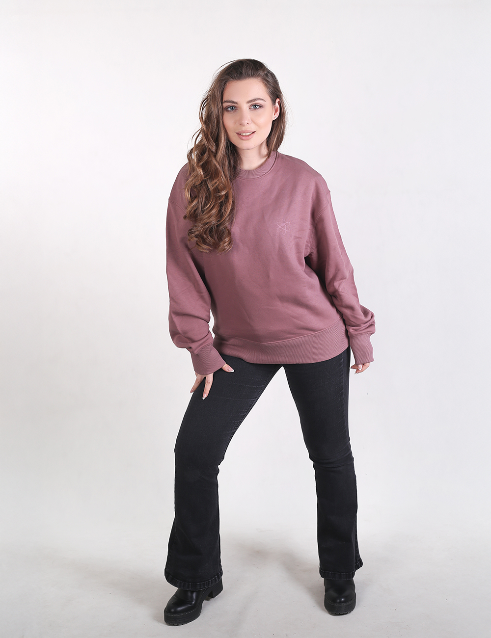 A woman stands against a plain white background, wearing a mauve crewneck sweatshirt, black flare jeans, and black chunky boots. She has long, wavy brown hair draped over one shoulder and is posing with one hand on her hip.