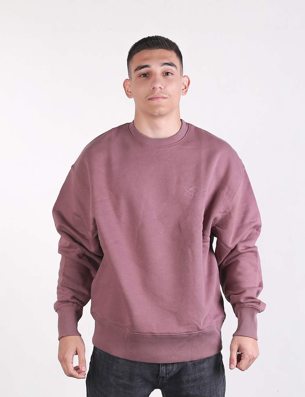 A young man stands facing forward against a plain, light grey background, wearing a mauve-colored oversized crewneck sweatshirt and dark charcoal or black jeans.