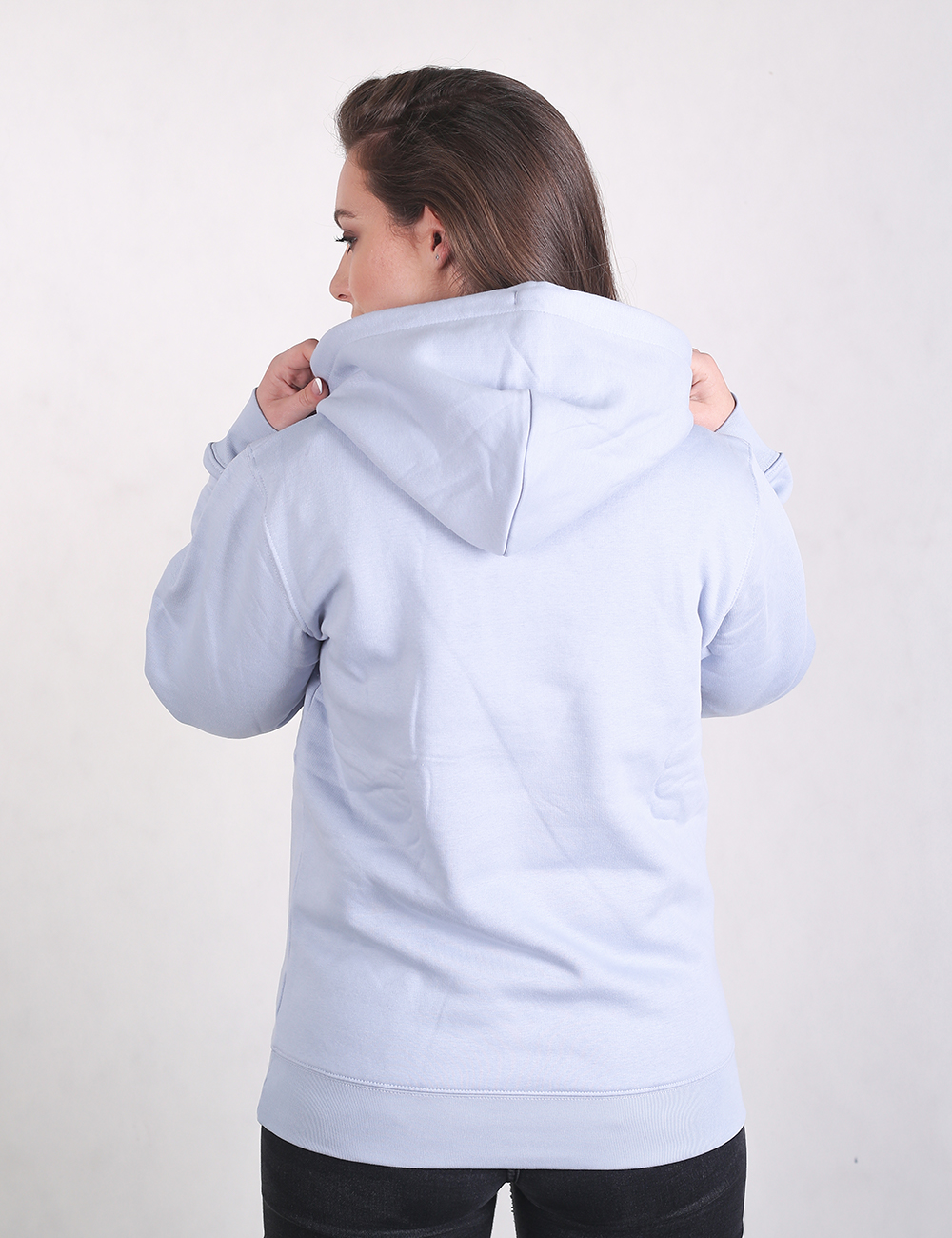 A person with long brown hair stands facing away from the camera, wearing a light blue, long-sleeved hooded sweatshirt and dark pants against a plain, light grey background.