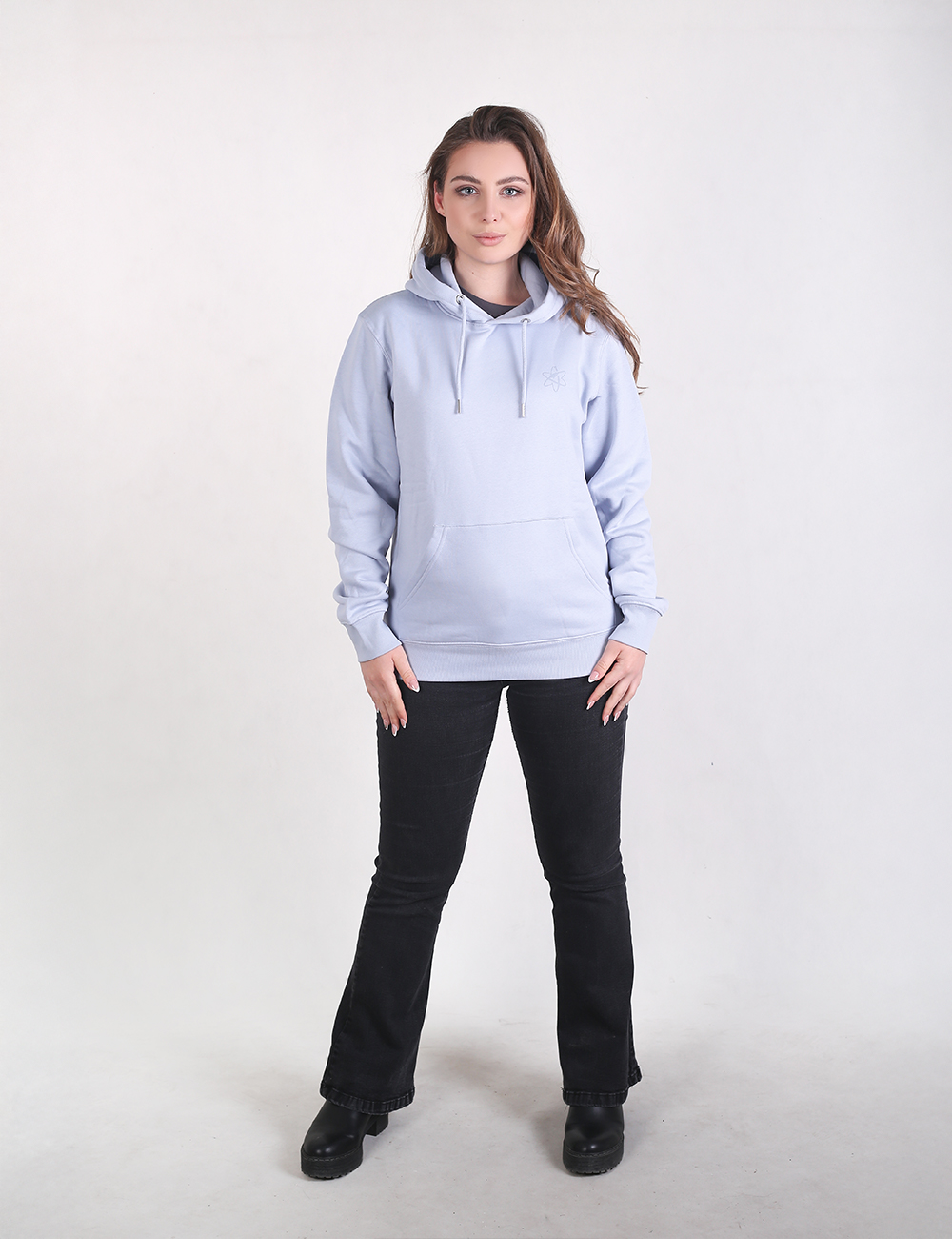 A woman stands against a plain white background wearing a light ice-blue hoodie with a front kangaroo pocket and drawstrings, paired with black flare pants and dark shoes. 