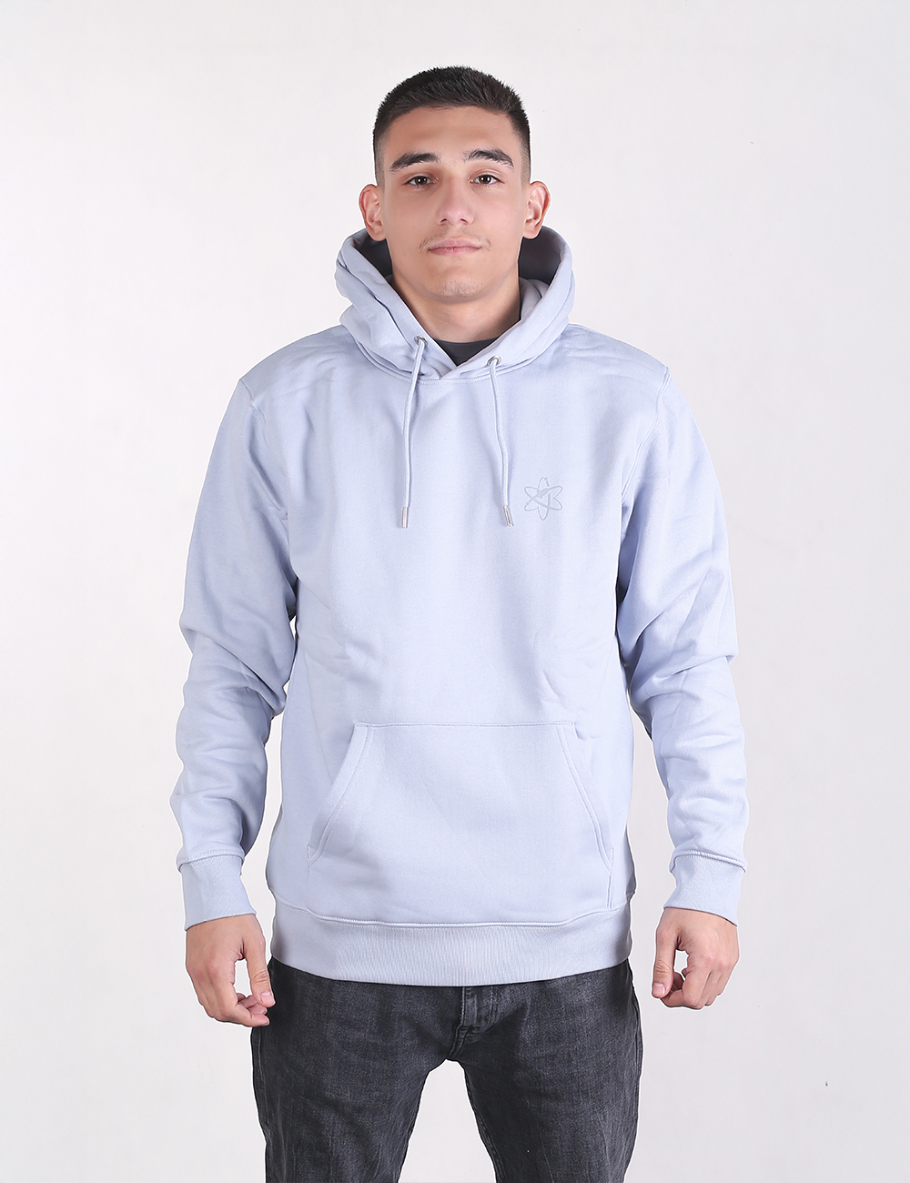 A young man with short dark hair stands looking forward against a plain white background, wearing a light sky-blue pullover hoodie with a front kangaroo pocket and dark charcoal jeans.