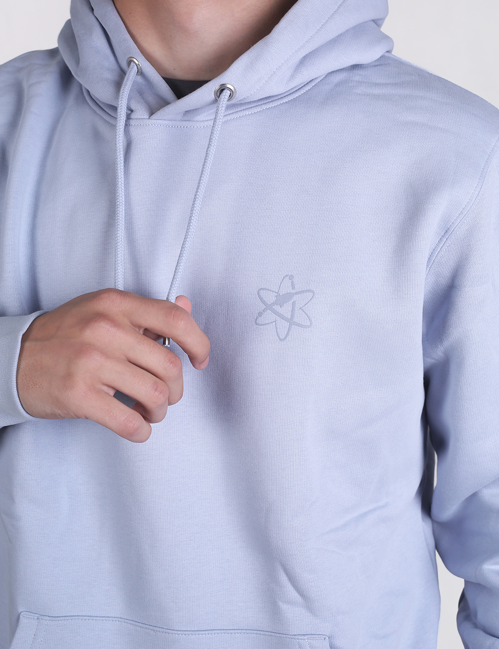 A close-up shot of a person wearing a light blue hoodie featuring a small, subtle atom symbol printed on the left chest. The person's left hand is visible, gently pulling on one of the hoodie's drawstrings.