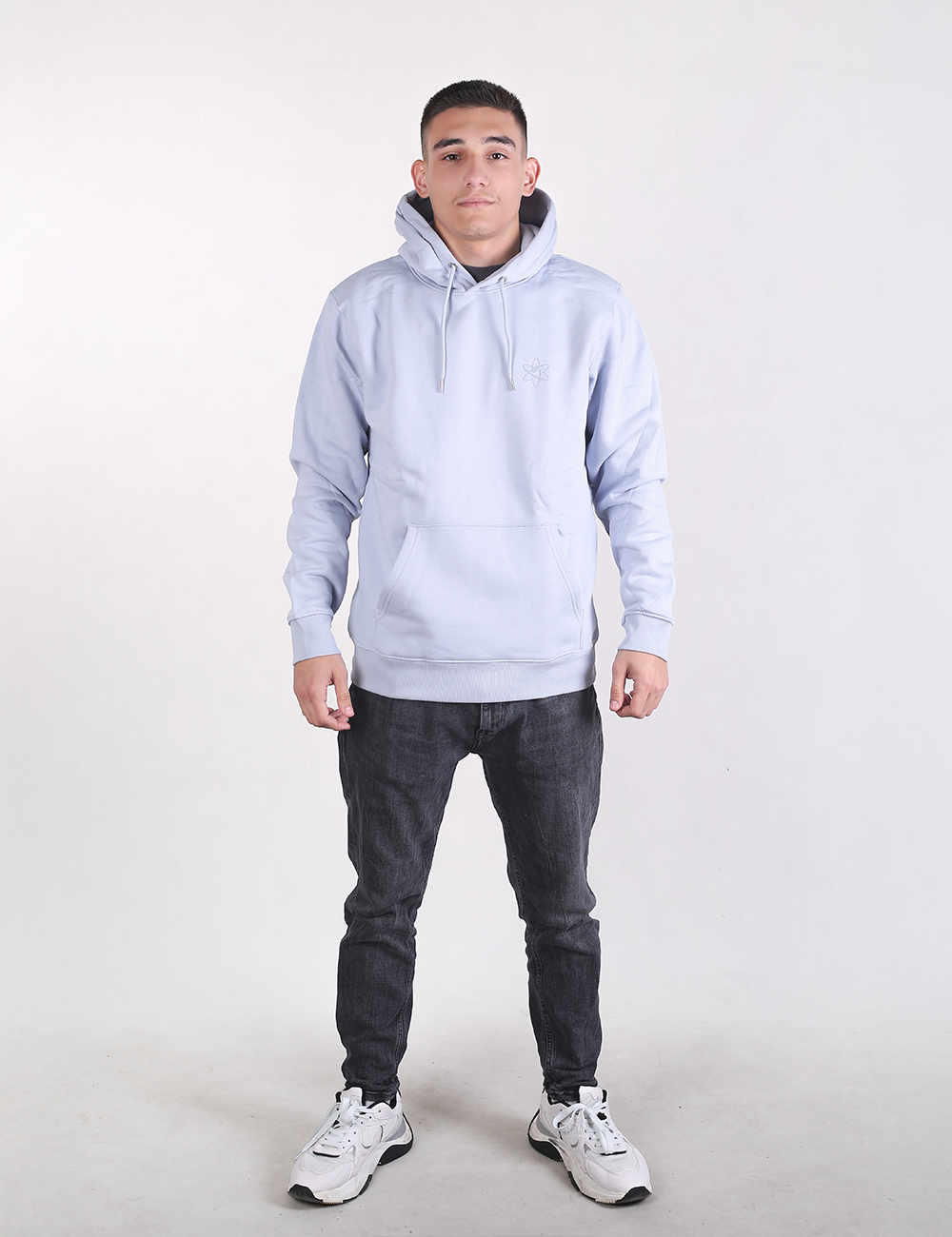 A man stands facing forward against a plain white background, wearing a light blue hoodie, dark grey slim-fit jeans, and white athletic sneakers.