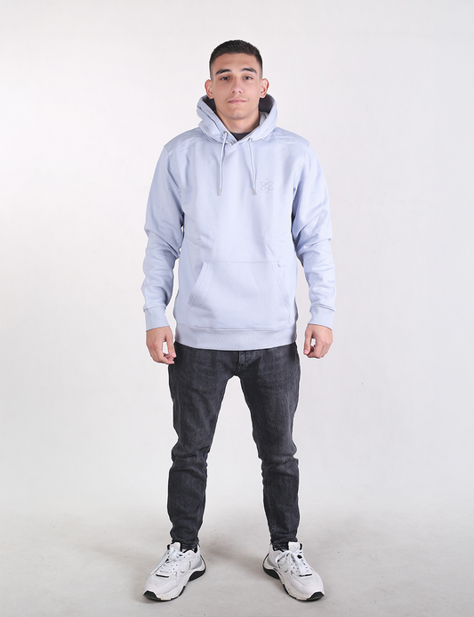 A man stands facing forward against a plain white background, wearing a light blue hoodie, dark grey slim-fit jeans, and white athletic sneakers.