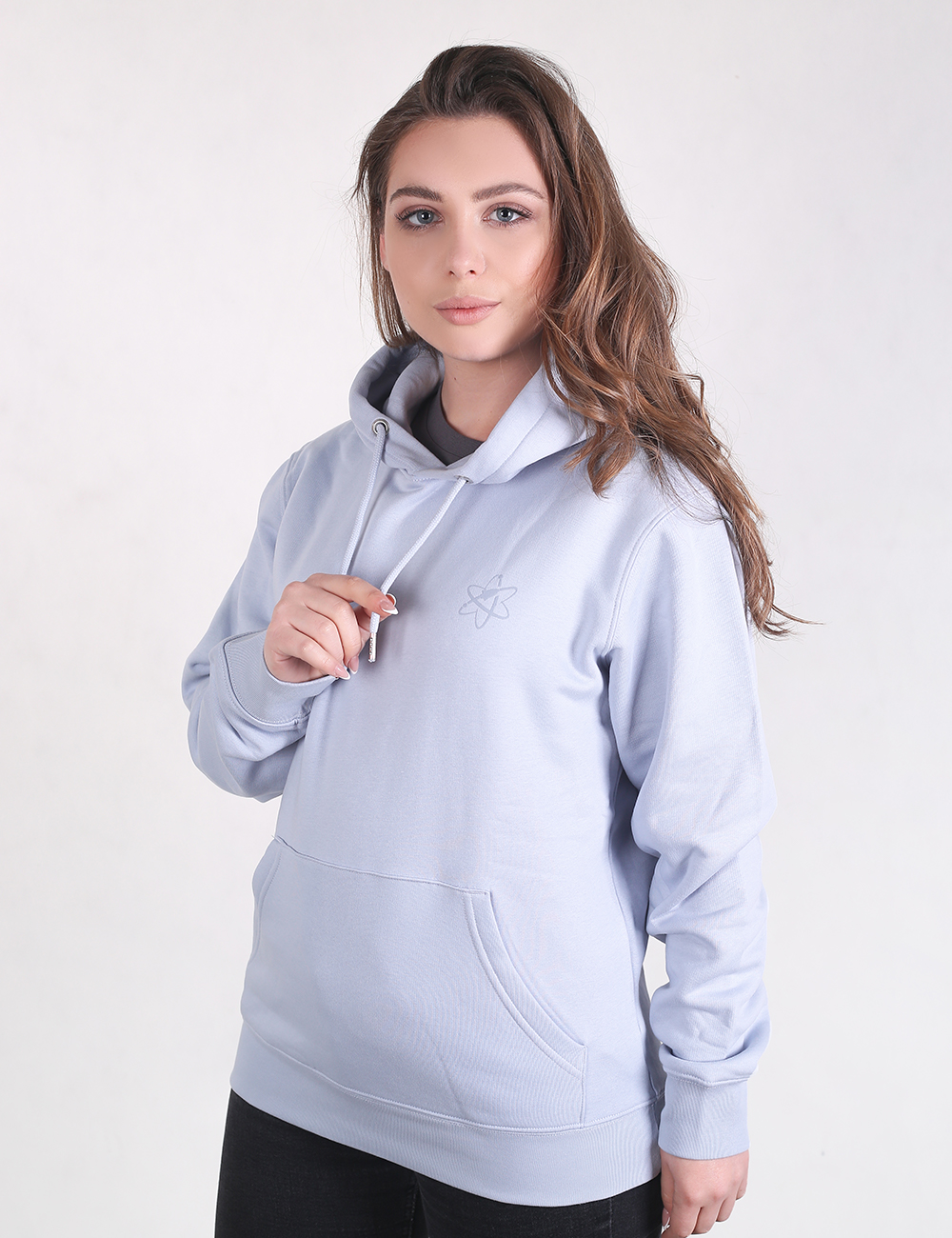  A young woman with long, light-brown wavy hair is pictured from the waist up against a plain white background, wearing a light blue pullover hoodie and black pants. Her right hand is slightly raised, holding one of the white drawstrings of her hood, while her left arm hangs relaxed at her side. The hoodie features a small, subtle embroidered logo on the upper left chest area and a spacious kangaroo pocket across the front waist.
