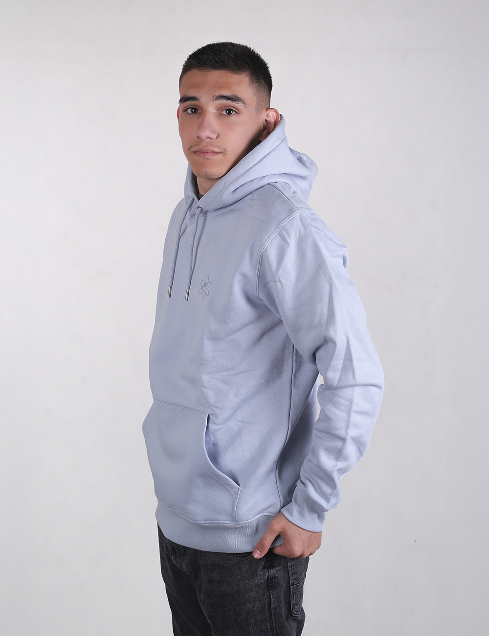A young man with short dark hair looking slightly over his shoulder while wearing a light blue or lavender drawstring pullover hoodie with a front kangaroo pocket and dark jeans, posed against a plain white background.