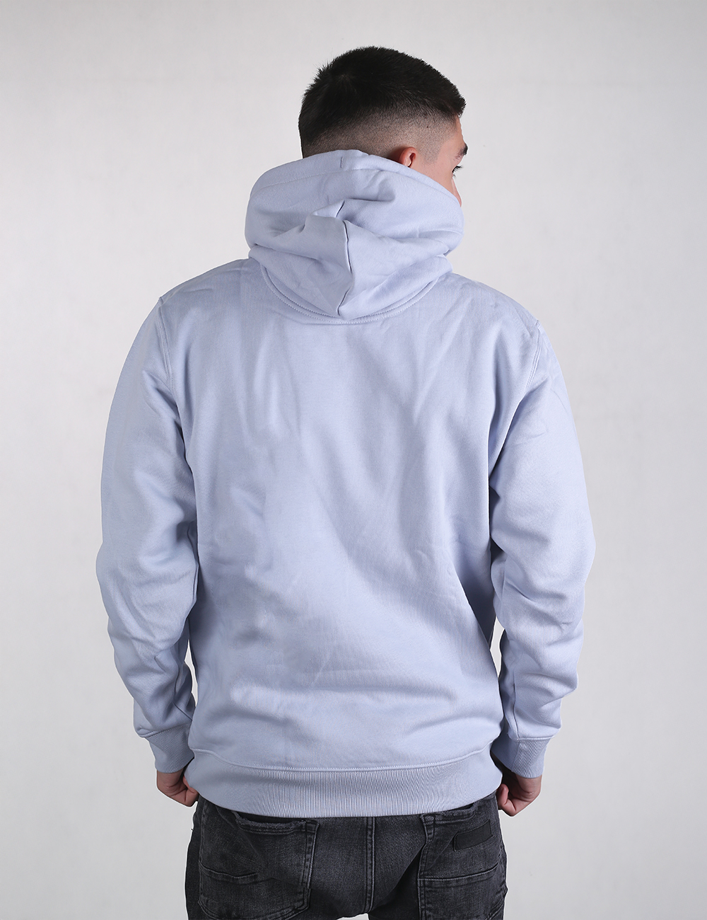 A young man is viewed from behind, wearing a light blue pullover hoodie and dark gray jeans. He has short dark hair with a faded cut on the sides. The hoodie features a structured hood pulled up loosely, ribbed cuffs at the wrists, and a ribbed hem at the waist.