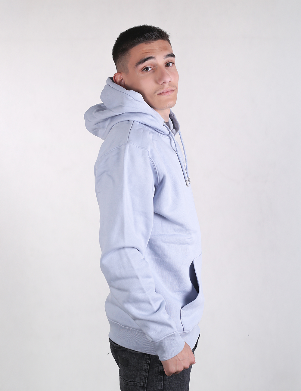 A young man with short dark hair, shaved closely on the sides, stands in a side-profile view against a plain, light grey background while looking over his shoulder toward the camera. He wears a light sky-blue pullover hoodie with a large hood and a front kangaroo pocket, paired with dark grey or black jeans.
