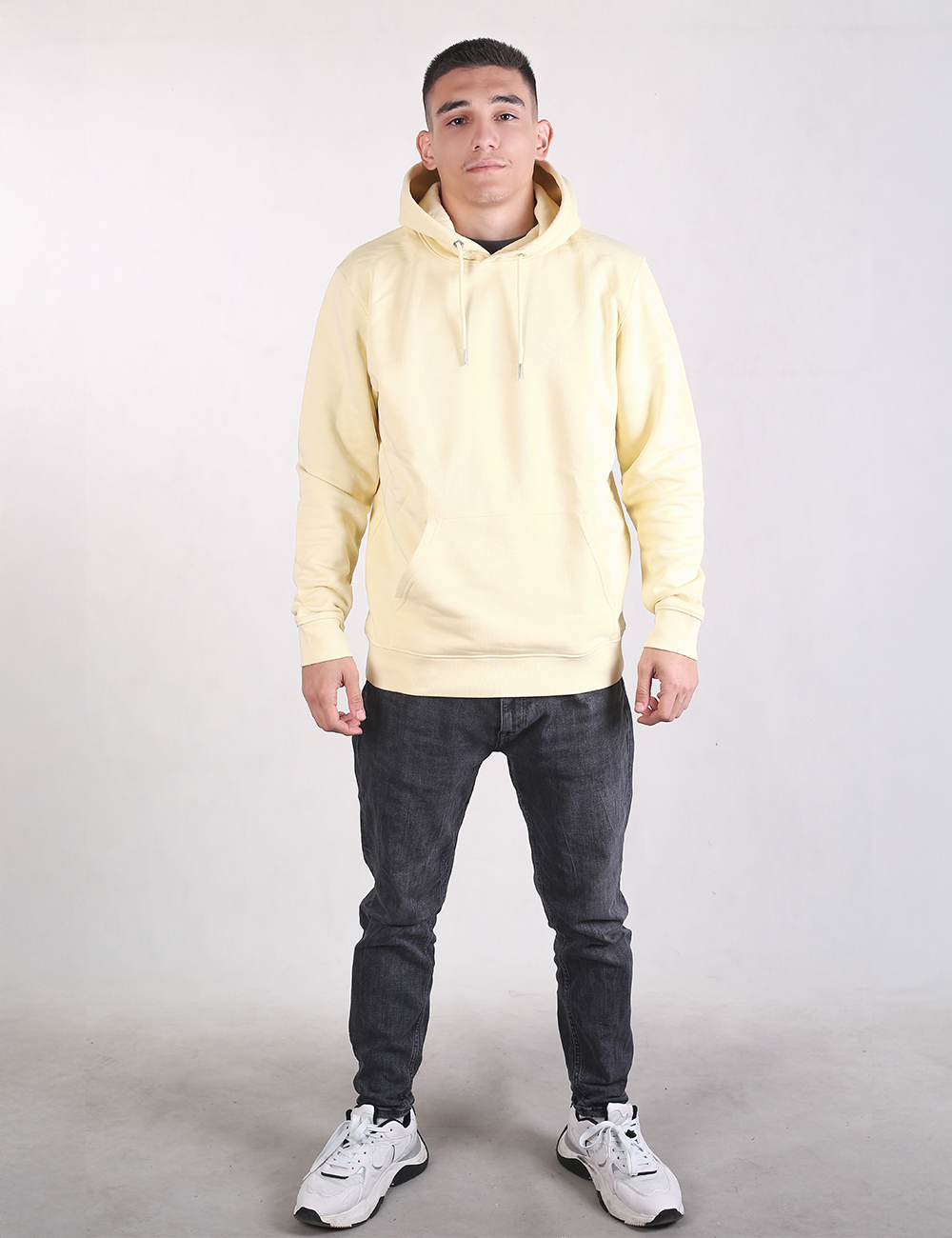 A full-length photo of a young man standing against a plain, light grey background. He is wearing a light yellow pullover hoodie, dark grey slim-fit jeans, and white athletic sneakers with chunky soles. He is facing forward with a neutral expression, arms relaxed at his sides, and feet spaced shoulder-width apart.