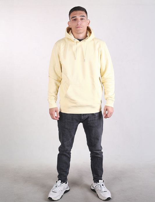 A full-length photo of a young man standing against a plain, light grey background. He is wearing a light yellow pullover hoodie, dark grey slim-fit jeans, and white athletic sneakers with chunky soles. He is facing forward with a neutral expression, arms relaxed at his sides, and feet spaced shoulder-width apart.