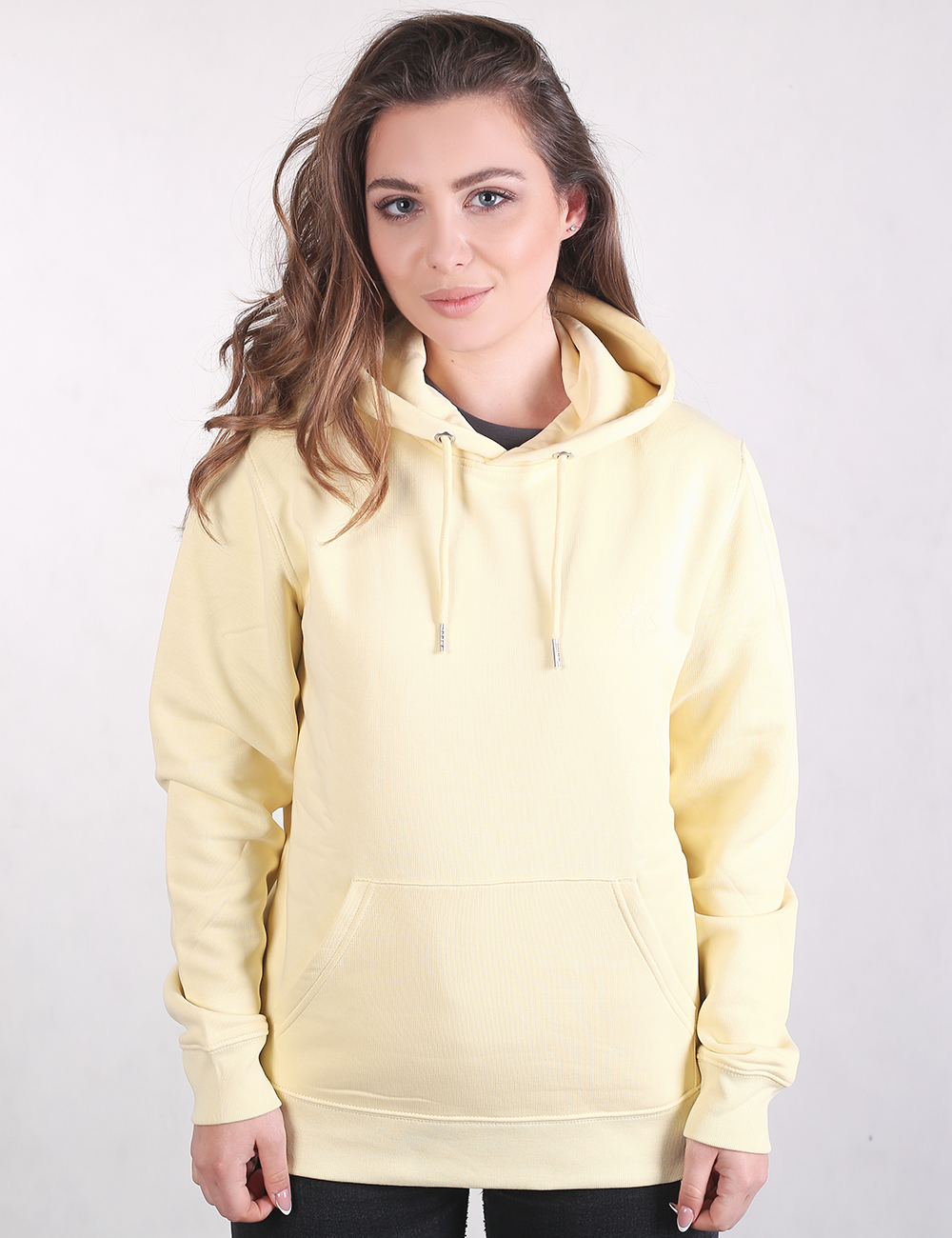 A woman with long, wavy light brown hair stands in a front-profile view against a plain light gray background. She is wearing a pale pastel yellow pullover hoodie with .