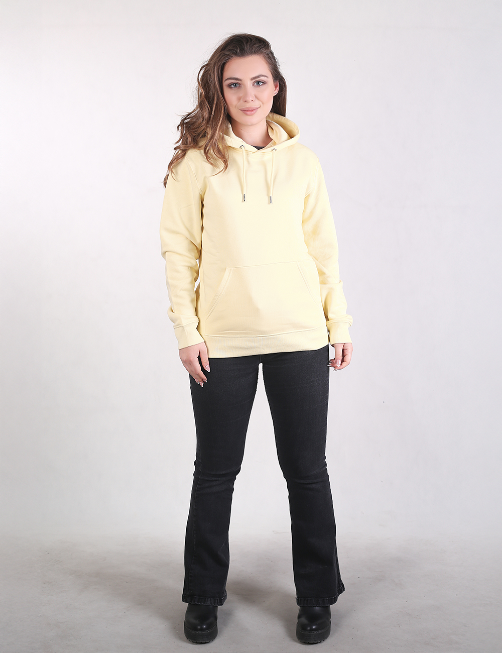 A woman with long, wavy light brown hair stands in a side-profile view against a plain light gray background. She is wearing a pale pastel yellow pullover hoodie with her right hand tucked into the front kangaroo pocket.