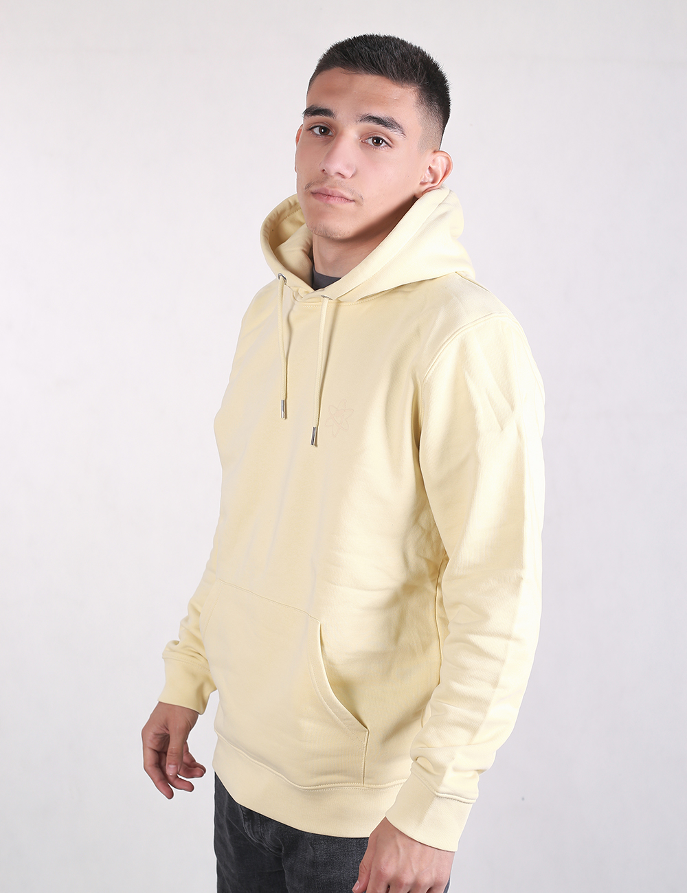 A young man with short dark hair standing in a side-profile view against a plain white background, wearing a light butter-yellow pullover drawstring hoodie and dark pants.