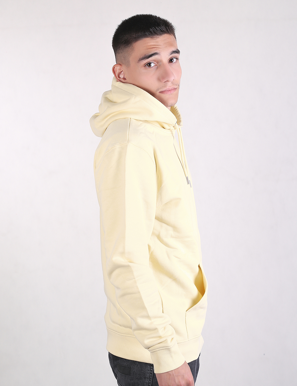 A young man with short dark hair looks over his shoulder toward the camera, captured from the side against a plain white background. He is wearing a light yellow pullover hoodie with a large front pocket and a drawstring hood, paired with dark jeans.