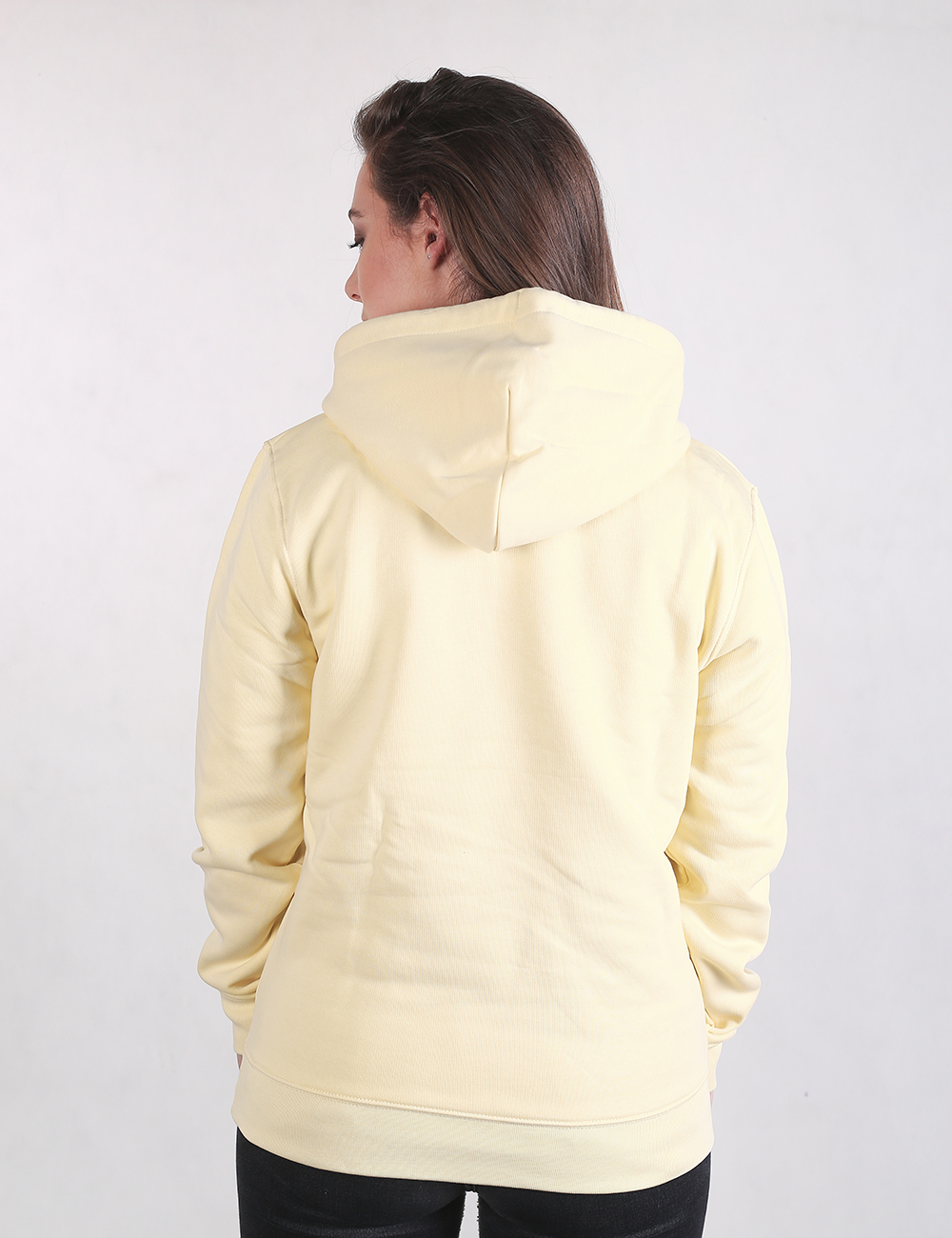 A woman with her back to the camera, wearing a light yellow pullover hoodie and dark pants against a plain white background.