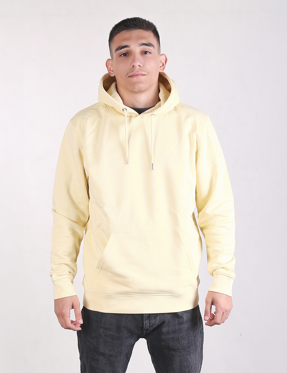 A young man with short dark hair stands looking forward, wearing a pale butter-yellow pullover hoodie with drawstring ties and a front kangaroo pocket, paired with dark grey or black jeans against a plain light background.