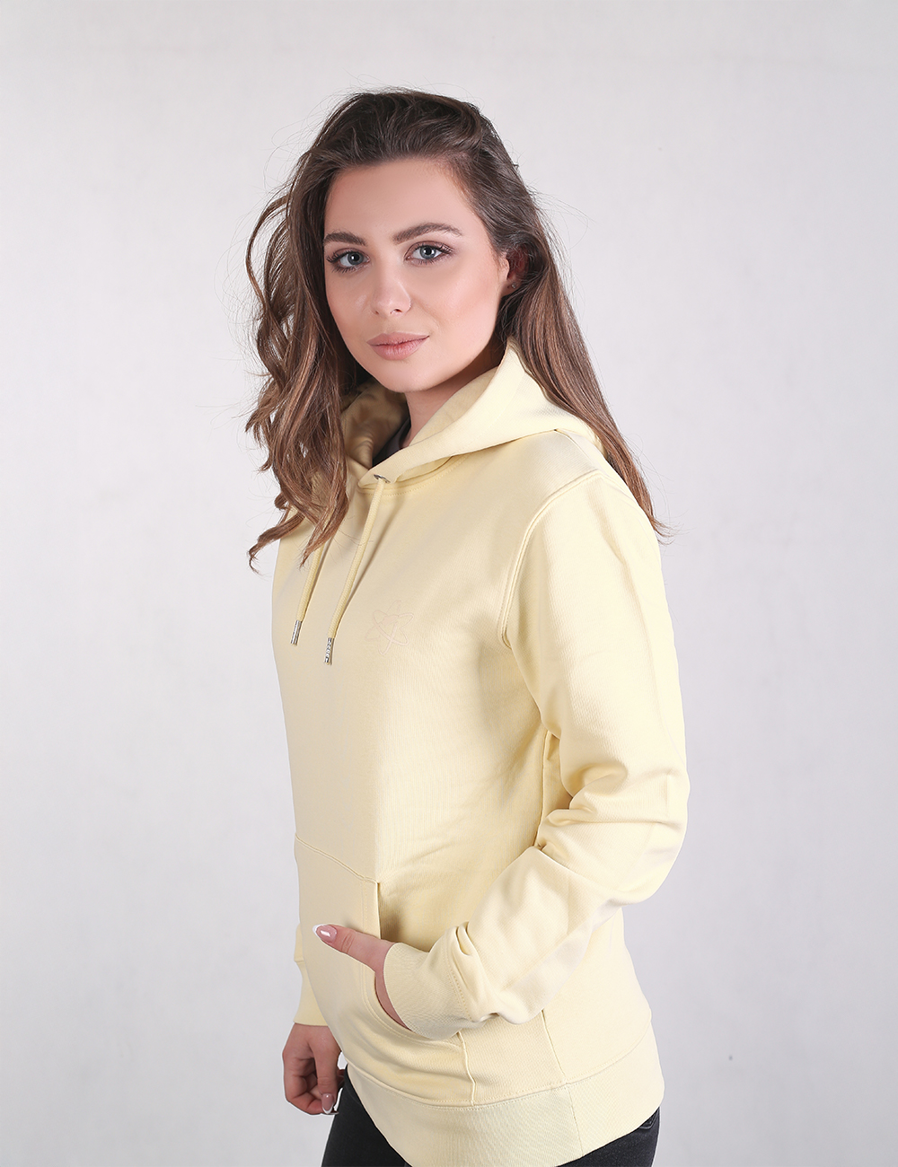 A woman with long, wavy light brown hair stands in a side-profile view against a plain light gray background. She is wearing a pale pastel yellow pullover hoodie with her right hand tucked into the front kangaroo pocket.