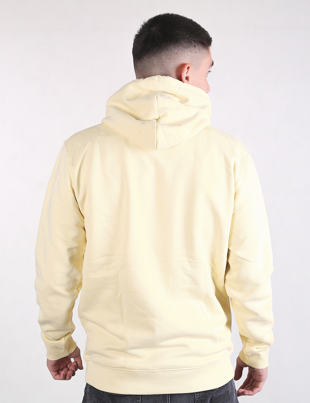 A young man with a short fade haircut is shown from the back, wearing a light pastel yellow pullover hoodie. The hoodie features a structured hood, dropped shoulders, and ribbed cuffs and hem. He is posed against a plain, light-colored background.