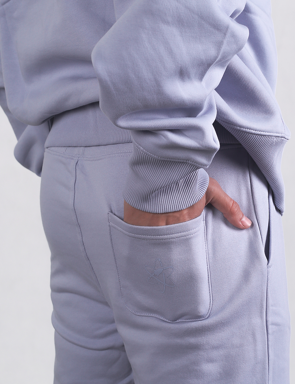 A close-up shot of a person from behind, wearing a light purple cotton fleece tracksuit. The individual has their hand tucked into the patch pocket on the upper right back of the joggers. The ribbed cuff of the matching lilac sweatshirt sleeve rests just above the pocket line.