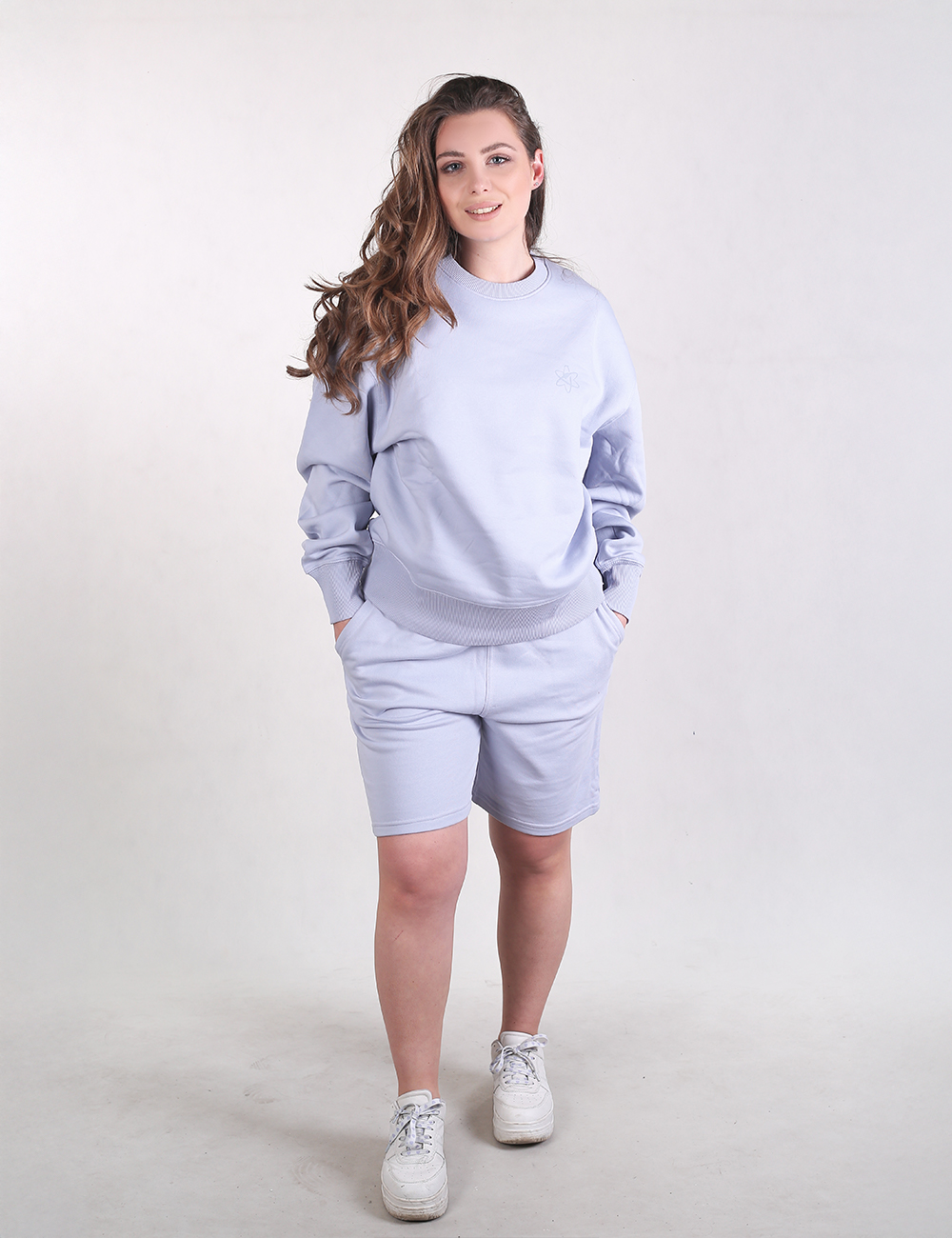 The image shows a pair of light grey-blue sweat shorts featuring a comfortable, casual design. They are mid-length, hitting a few inches above the knee, and feature a relaxed fit with deep side pockets where the wearer's hands are placed. The fabric appears to be a thick, soft sweatshirt material matching the top.