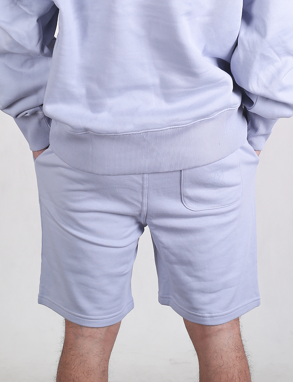  The person has their hands in the front pockets of a plush, relaxed-fit cotton sweatshirt featuring a thick ribbed hem. Below, they are wearing matching mid-thigh length sweat shorts that feature a single open pouch pocket on the right rear side.