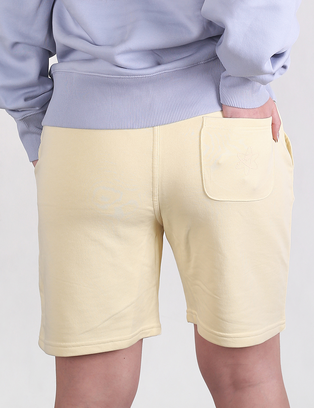 A rear view of a person wearing pale yellow sweat shorts paired with a light lavender or periwinkle sweatshirt. The person's left hand is partially tucked into the single patch pocket on the upper right side of the shorts. The shorts have a casual, cotton fleece texture with raw or stitched hems, and the sweatshirt features a ribbed waistband and cuffs.
