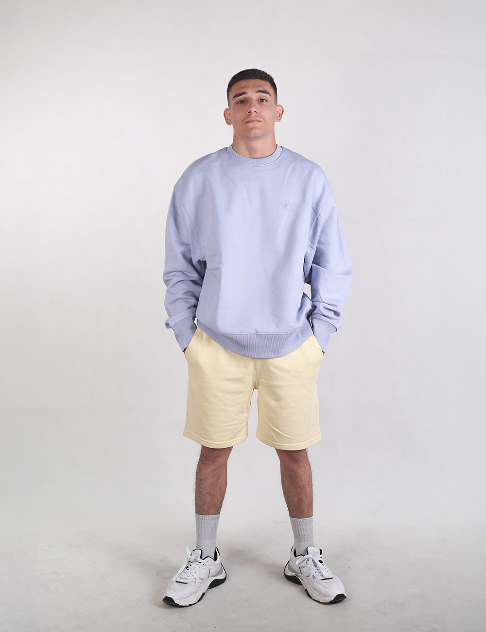 A young man with short dark hair stands looking forward against a plain white background, wearing a baggy, light blue crewneck sweatshirt and pale, butter-yellow sweat shorts. His hands are tucked casually into his shorts pockets.
