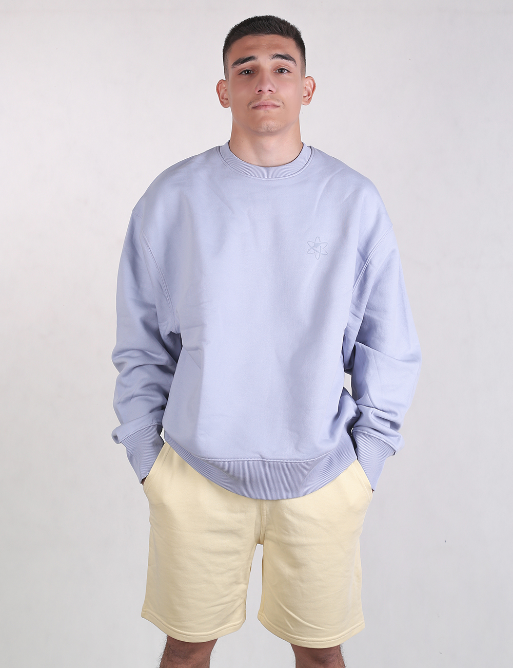 A young man with short dark hair stands looking forward against a plain white background, wearing a baggy, light blue crewneck sweatshirt and pale, butter-yellow sweat shorts. His hands are tucked casually into his shorts pockets.
