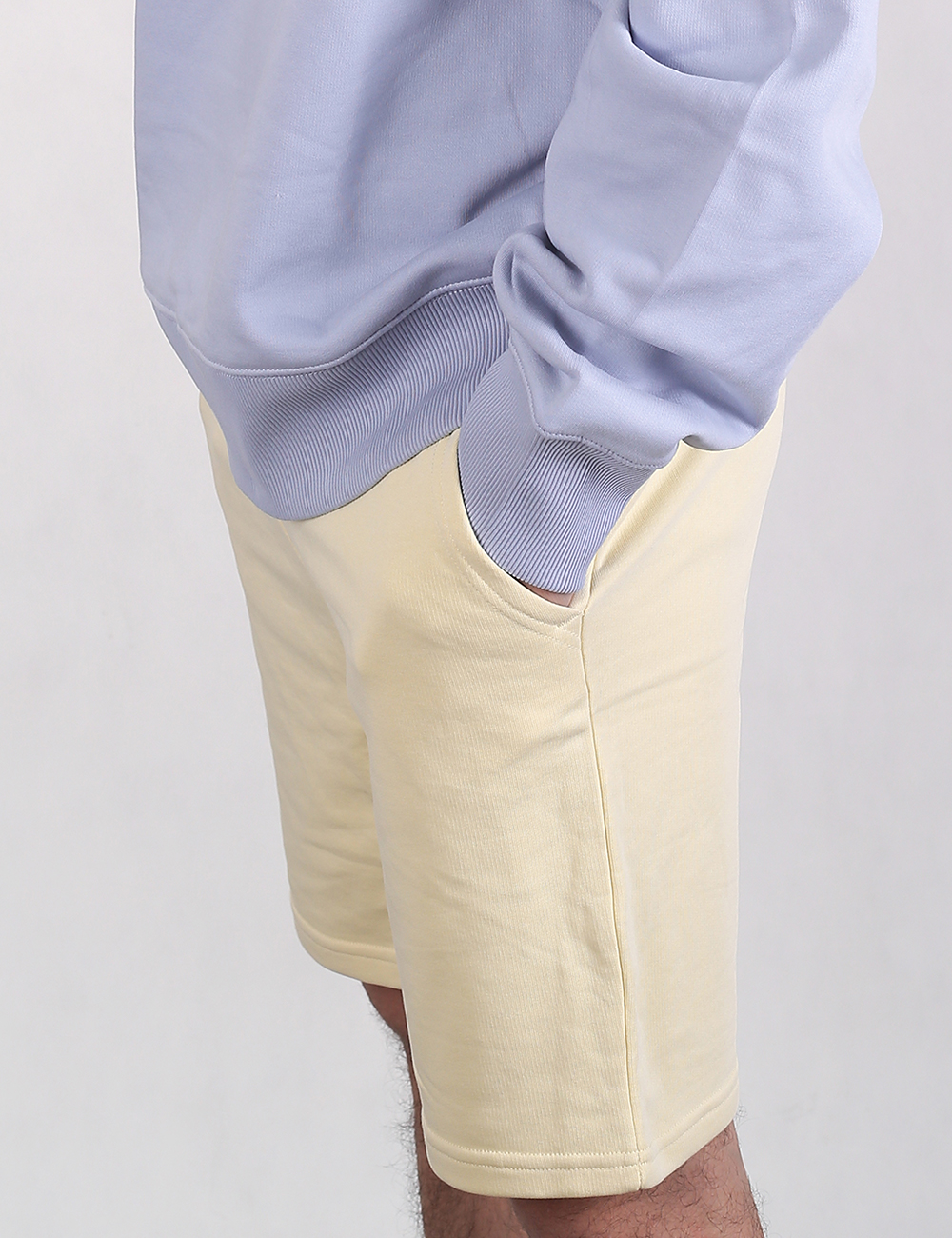A side-angle, close-up view of a person wearing a pale blue long-sleeve sweatshirt and light cream-yellow sweat shorts. The individual has their right hand resting in the side pocket of the shorts.