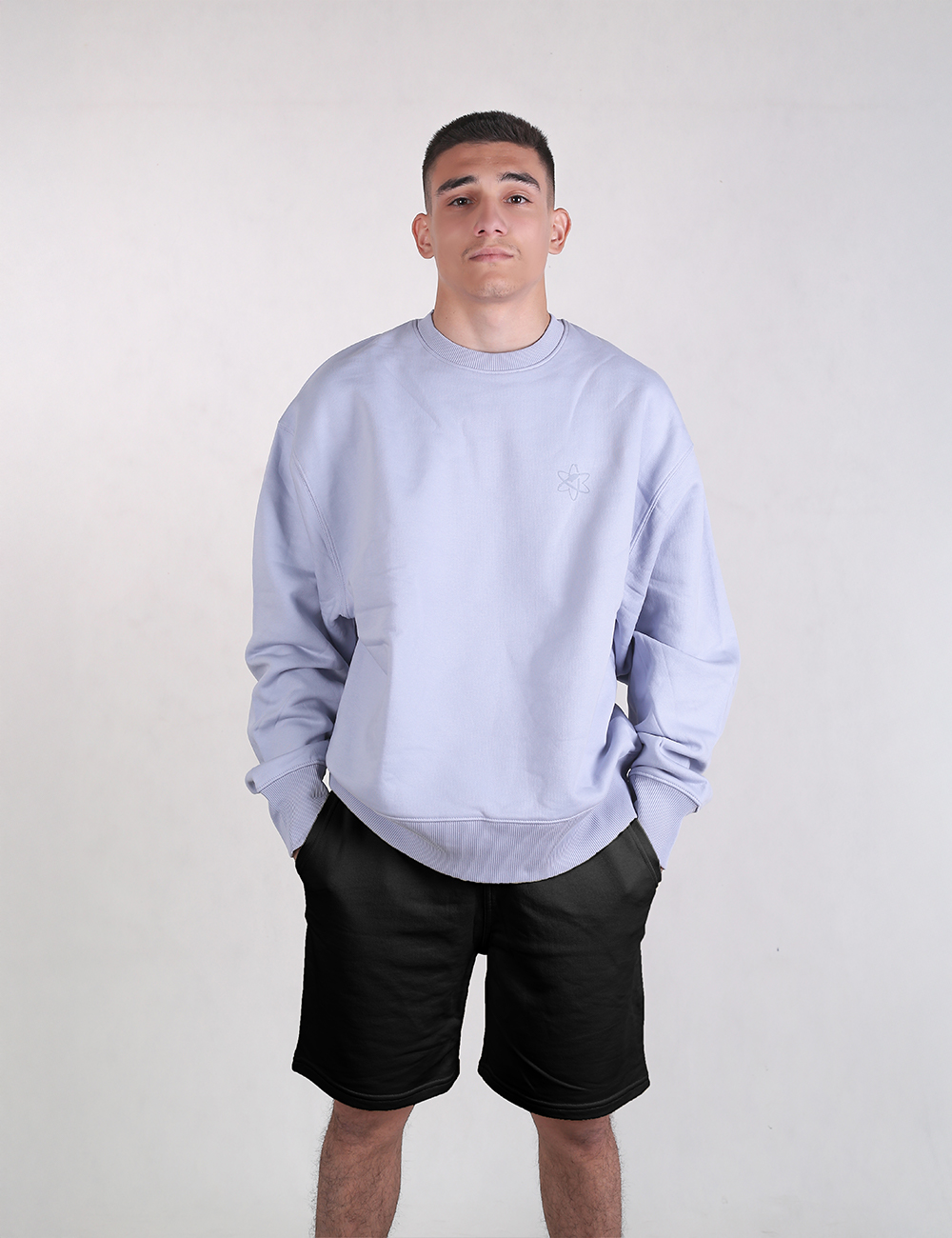 A young man stands facing forward against a plain light gray background. His hands are tucked into the pockets of his black athletic shorts. 
