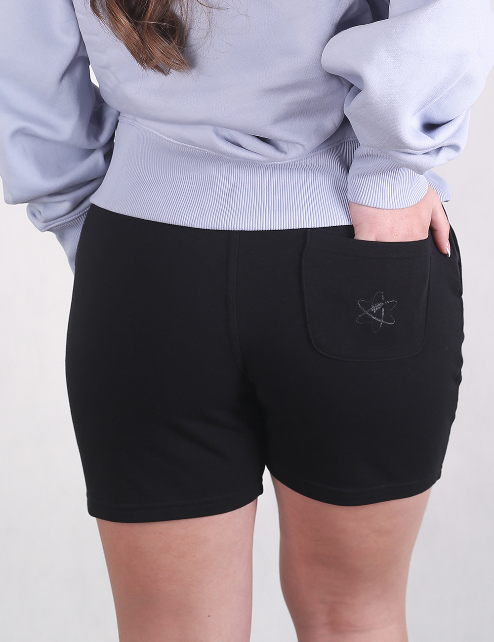 A person is shown from the back wearing a light lavender or light blue long-sleeved sweatshirt and black fitted shorts. Their left hand is resting inside the back pocket of the shorts, which features a subtle, dark embroidered atom or rocket-themed logo.