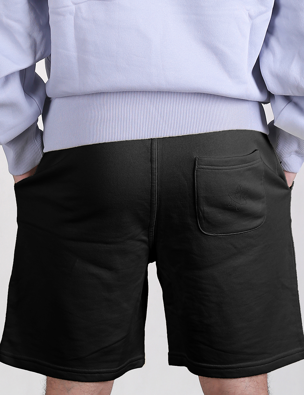 Rear view of a person wearing black sweatshirt shorts paired with a light blue or lavender hoodie, showcasing a single patch pocket on the right side of the shorts.