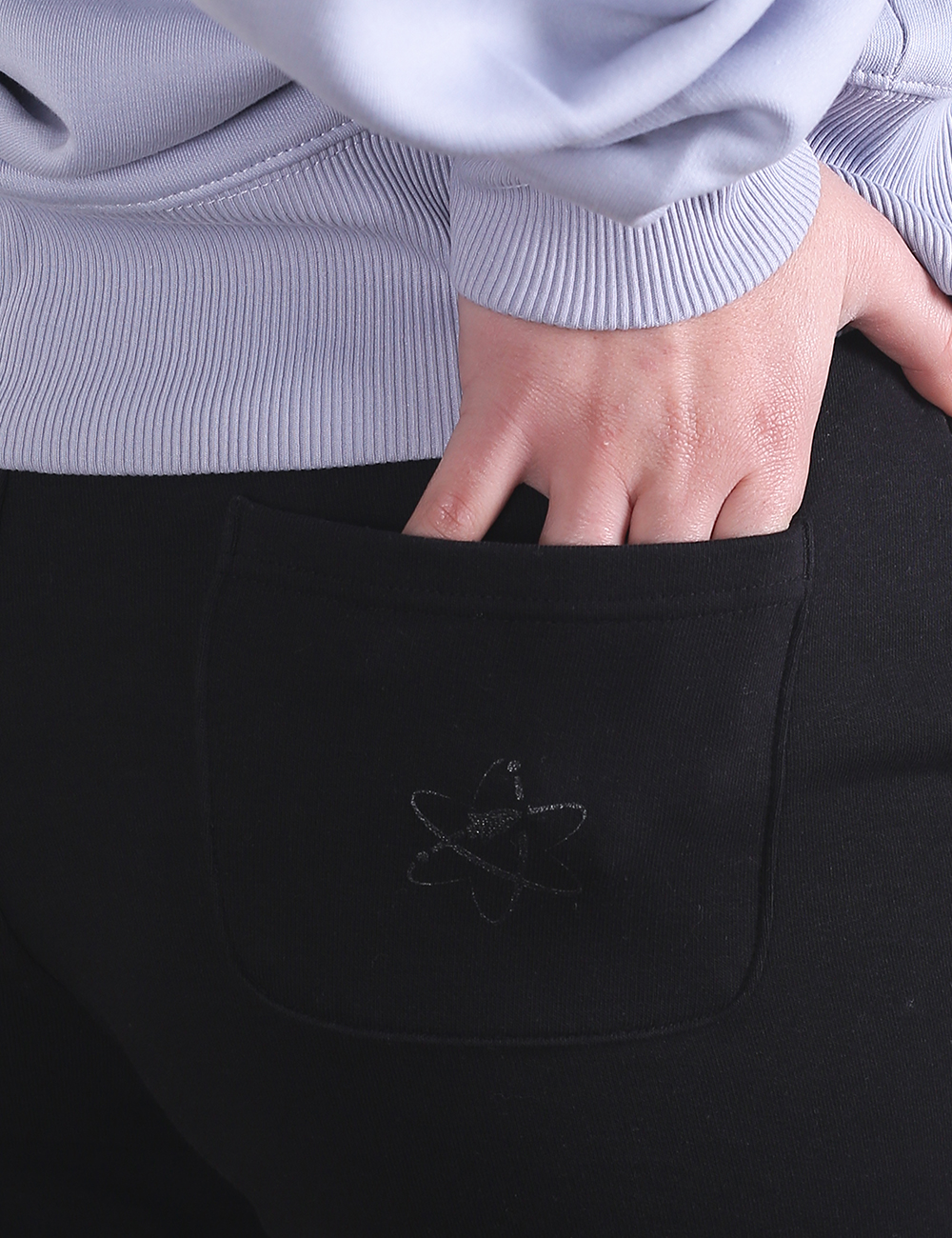 A close-up photograph focuses on a person's hand sliding into the back pocket of black pants. The hand enters from above, with fingers partially submerged into the pocket opening.On the center of the pocket, a subtle black embroidered logo depicts intersecting atomic or planetary orbital rings. Above the pants, the ribbed hem of a light lavender or periwinkle blue sweatshirt is visible.
