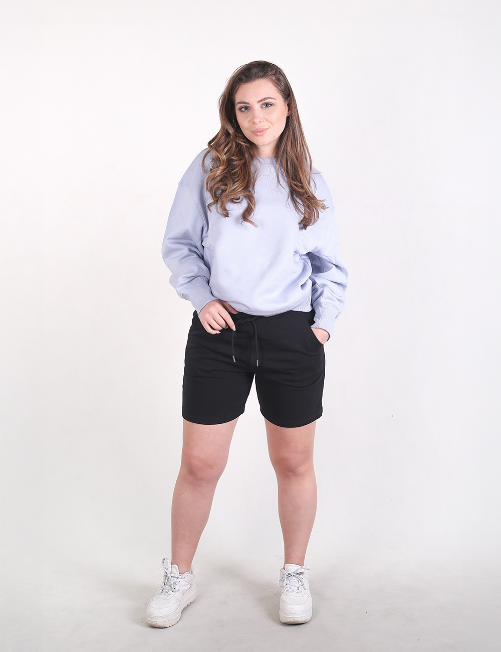 A young woman wearing a light lavender or light blue long-sleeve sweatshirt and black athletic drawstring shorts. Their left hand is resting inside the left side pocket of the shorts, while their right hand holds onto the waistband near the tied drawstring cord.