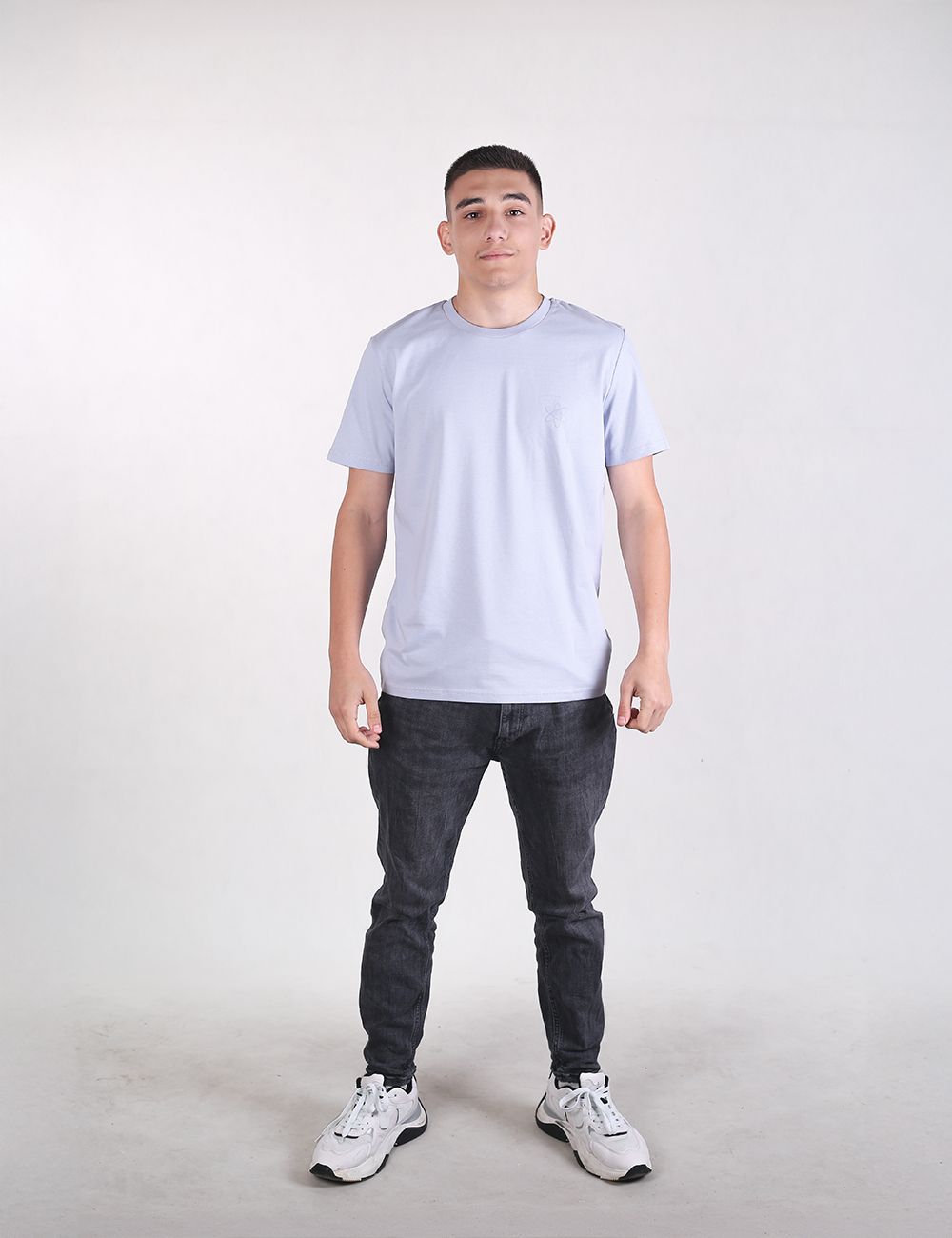 A man with short dark hair stands facing forward against a plain, light grey background, wearing a light blue  t-shirt, dark charcoal slim-fit jeans, and white athletic sneakers with black accents.