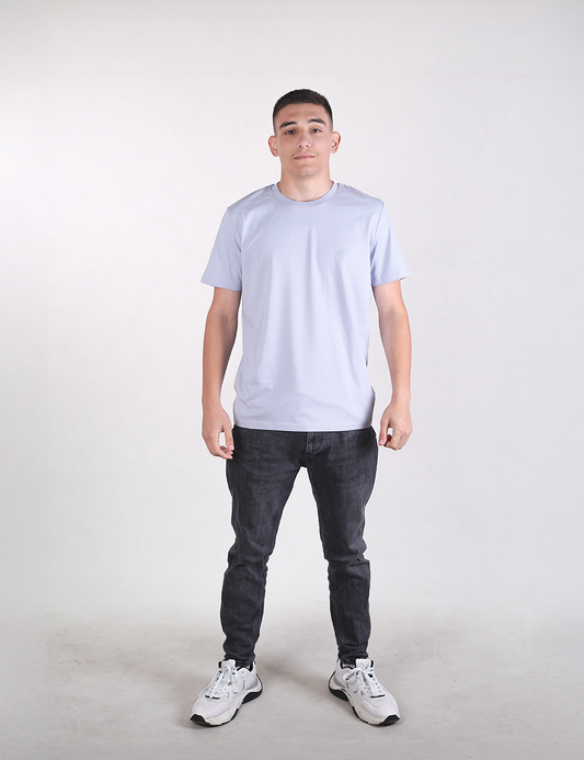 A man with short dark hair stands facing forward against a plain, light grey background, wearing a light blue  t-shirt, dark charcoal slim-fit jeans, and white athletic sneakers with black accents.
