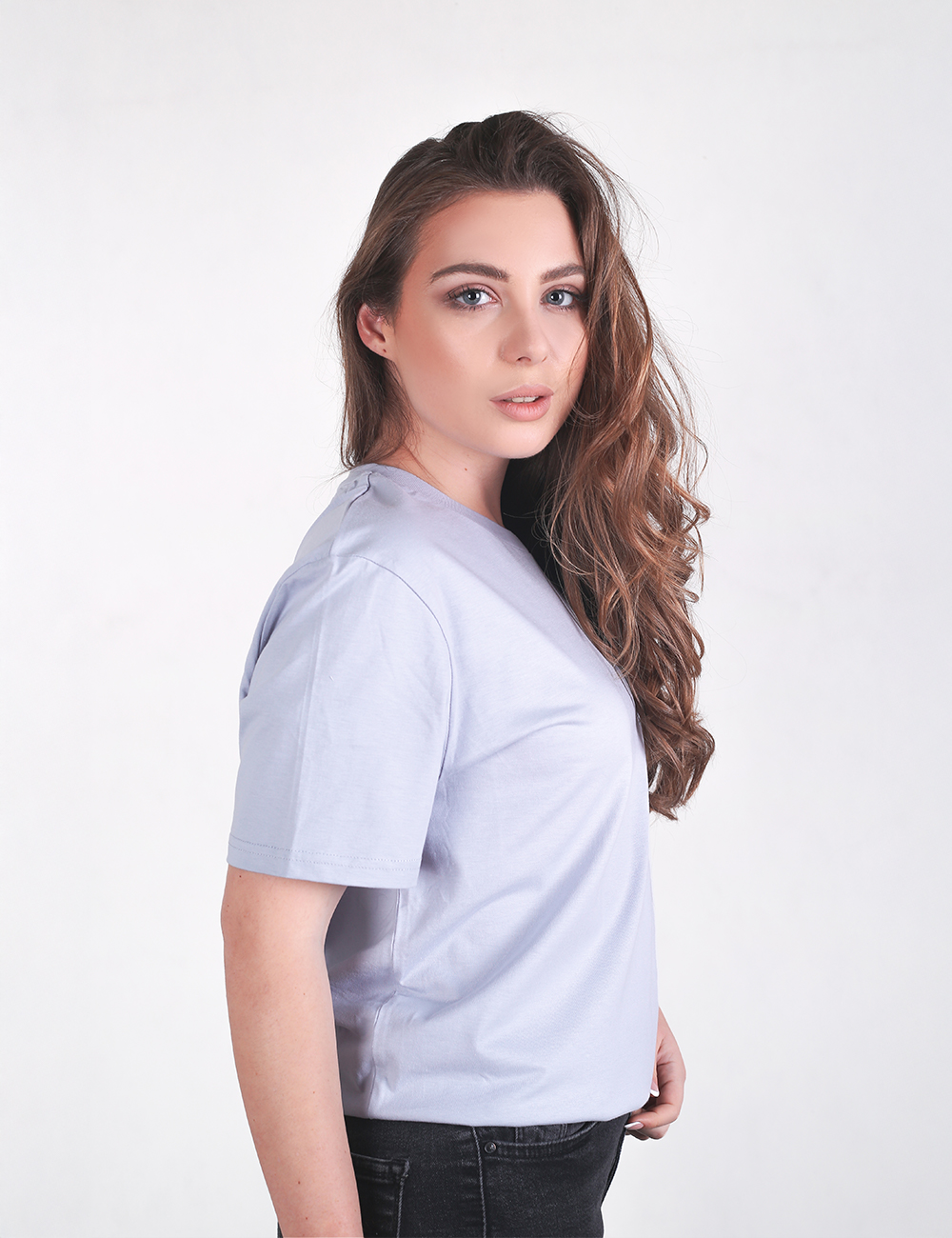 A young woman with long, wavy brown hair is standing in a side-profile pose against a plain, off-white background. She is wearing a plain, light-grey crew neck t-shirt and black jeans. 