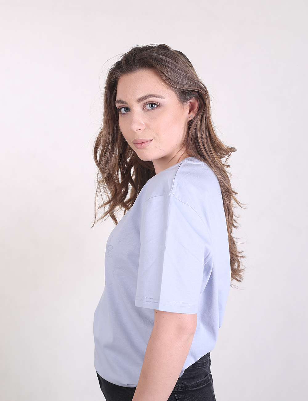 A young woman with long, wavy brown hair is standing in a side-profile pose against a plain, off-white background. She is wearing a plain, light-grey crew neck t-shirt and black jeans. 